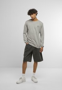 Cămașă gri cu mânecă lungă și un mic logo negru, asociată cu pantaloni scurți din denim închisi la culoare. Modelul poartă adidași albi și șosete, stând în fața unui fundal neutru.