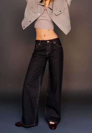 Persona snella che indossa jeans neri a gamba larga, top grigio corto, giacca grigia sollevata e scarpe rosso scuro, posando contro uno sfondo semplice.