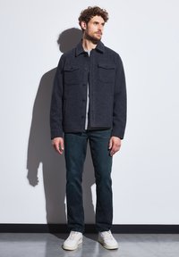 Tmavě šedý vlněný kabát s velkými předními kapsami, zapínáním na knoflíky a límcem. Nosený přes světle šedou košili, spojený s tmavými džíny a bílými teniskami.