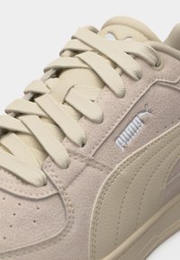 Κοντινή λήψη μπεζ αθλητικού παπουτσιού Puma με σουέτ και δερμάτινες υφές, κορδόνια σε κρεμ απόχρωση και ορατό λογότυπο Puma στην πλαϊνή επιφάνεια.