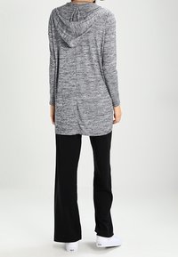 Dlouhá šedá mikina s kapucí vyrobená z texturované látky, doplněná černými rozšířenými kalhotami. Mikina má kapuci se stahovací šňůrkou a mírně delší zadní stranu.