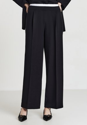 Femme portant un pantalon large noir avec une taille élastique blanche et des chaussures noires à bout pointu, se tenant avec les mains dans les poches.