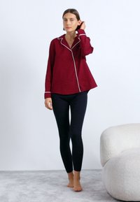 Burgundská košeľa s gombíkmi a bielym pásikovaním, zakrivený spodný okraj, v kombinácii s čiernymi legínami. Model stojí na svetlošedej koberci. Žiadne výrazné vzory.