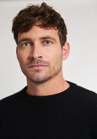 Maglione nero lavorato a maglia con colletto a coste, indossato da un uomo con capelli corti e ricci e una barba sottile. Tessuto testurizzato, design aderente.