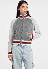 Maglione grigio e crema con zip e dettagli a coste in rosso. Presenta un colletto alto e tasche laterali, con una vestibilità rilassata e maniche a contrasto.
