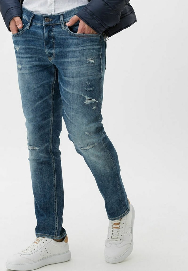 Vaqueros de denim azul ajustados con detalles desgastados, diseño de cinco bolsillos y zonas desvaídas. Combinados con zapatillas blancas, mostrando un look casual.