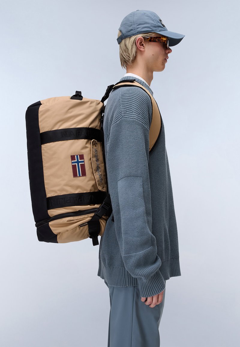 Beige and black padded backpack with a Norwegian flag patch, featuring straps, zippers, and a side handle, worn by a person in a grey sweater.