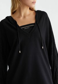 Schwarzer Hoodie mit V-Ausschnitt, ausgestattet mit Ärmeln im Spitzenmuster. Die Kordeln haben Metallspitzen und eine gebrandete Innenschicht ist sichtbar.