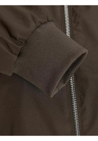 Bruine bomberjack mouw met ribbelstructuur, voorzien van een metalen rits en geplooide stof op de schouder, wat een slank ontwerp benadrukt.