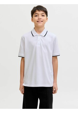 Chlapec stojí a usmívá se, má na sobě bílé polo tričko s černým lemem a černé kalhoty, na jednobarevném světle pozadí.