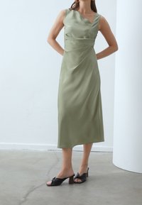 Robe midi en satin vert olive avec une épaule dénudée, des détails drapés à la taille et une texture lisse ; assortie à des mules noires à talons.