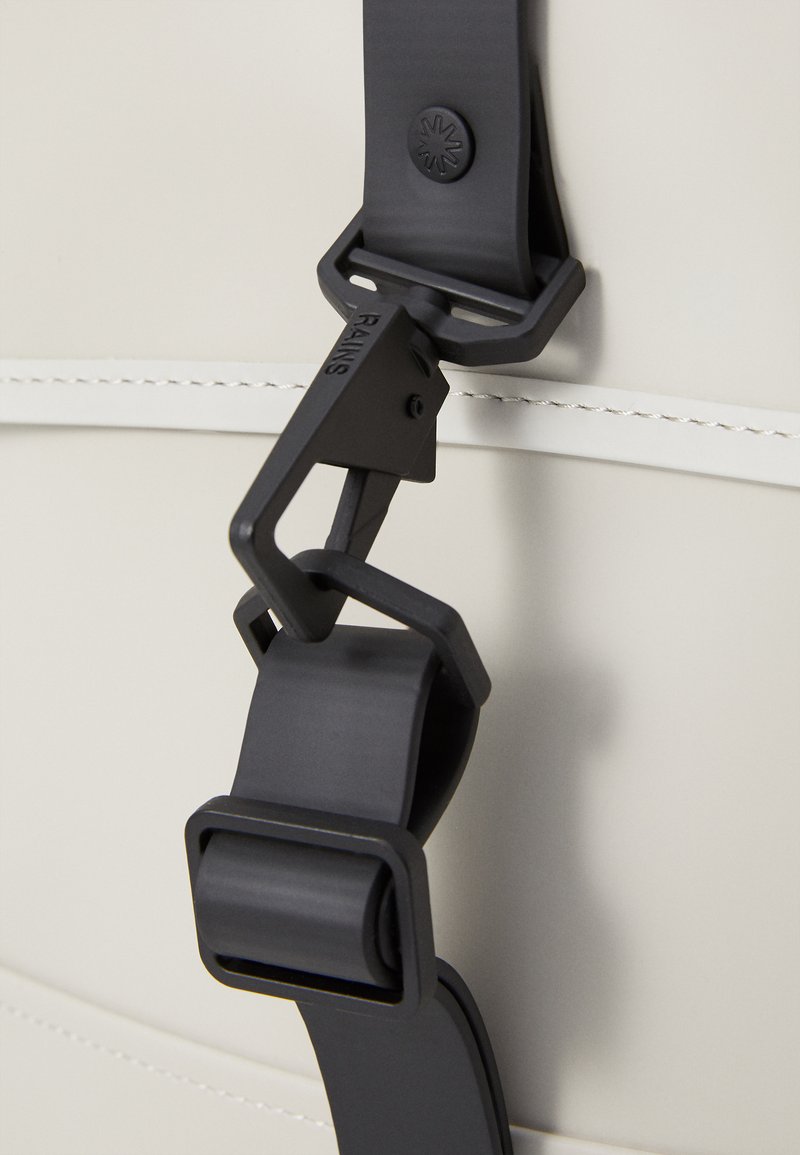 Boucle de plastique noir attachée à une sangle de couleur claire sur un sac. Paroi texturée avec un subtil détail de couture visible à l'arrière-plan.