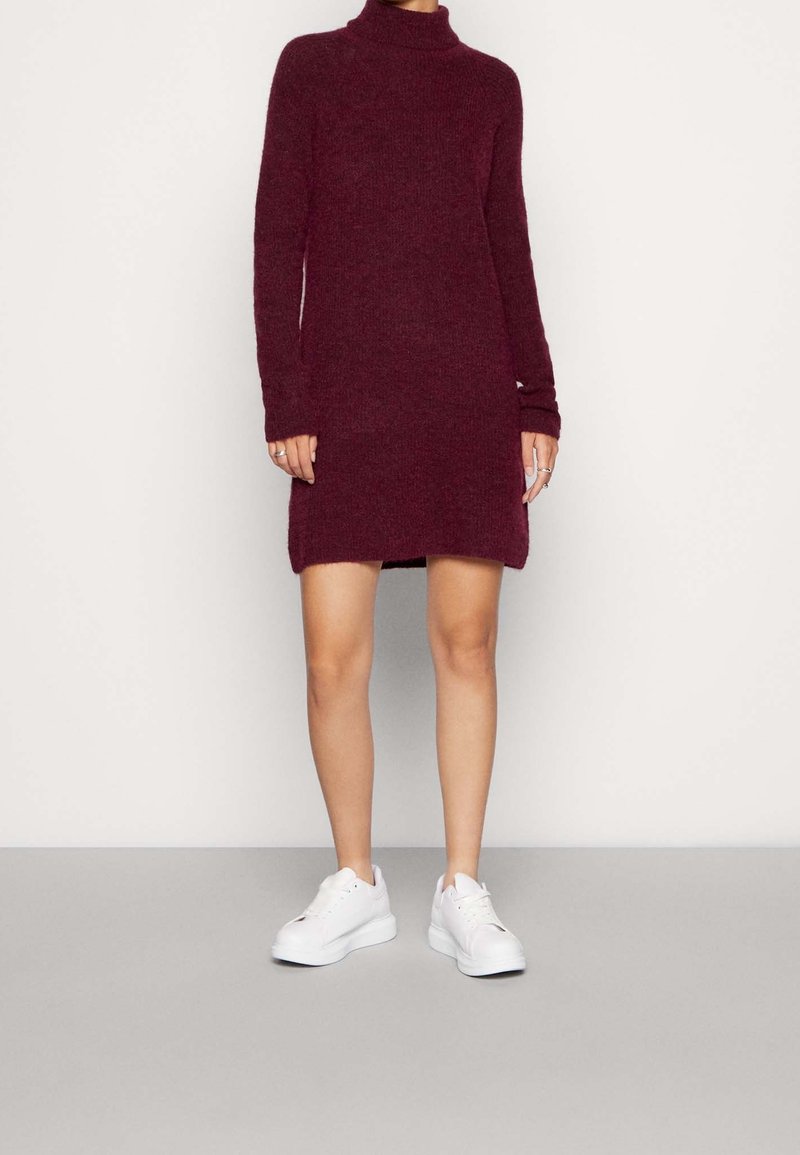 Burgunderfärgad stickad turtleneck-klänning med långa ärmar, med rak passform och sidslitsar, som bärs med vita sneakers.