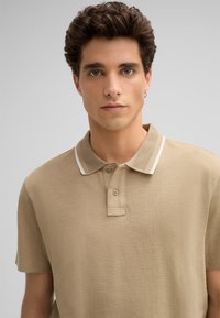 Beige Poloshirt mit strukturiertem Stoff, zwei-Knopf-Blende und weißem Kontrastkragen. Kurze Ärmel und lockere Passform. Keine sichtbaren Muster.