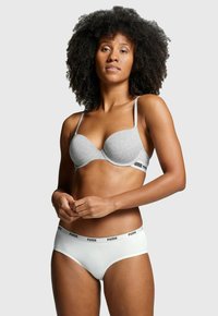 Frau mit lockigem Haar, die einen grauen BH und weiße Unterwäsche mit Puma-Logo auf einem schlicht hellen Hintergrund trägt.