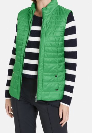 Woman wearing a green quilted vest over a black and white striped long-sleeve sweater and black pants, standing against a white background.