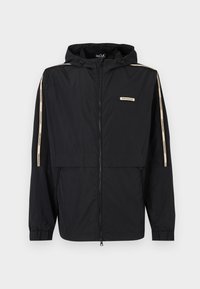 Chaqueta rompevientos negra con capucha, con cremallera frontal, puños elásticos y franjas laterales beige. Tela ligera y lisa.