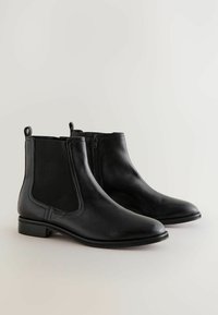Botas negras de cuero hasta el tobillo con paneles laterales elásticos y una cremallera, exhibidas sobre un fondo neutro.