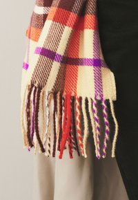 Écharpe en laine à carreaux colorée avec extrémités à franges dans des nuances de marron, orange, crème et violet, drapée sur un tissu noir et beige.
