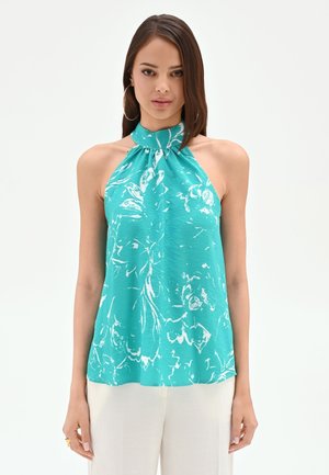 Femme portant un haut dos nu turquoise avec des motifs floraux blancs et un pantalon blanc, debout devant un fond clair uni.