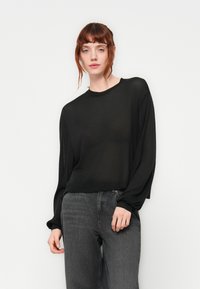 Haut noir transparent à manches longues avec un col rond et une coupe loose. Porté avec un jean gris taille haute. Texture douce avec un léger tombé.
