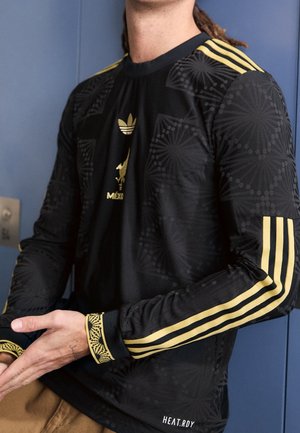 Person iført en sort langærmet Adidas-trøje med guldstriber, guld Mexico-logo og geometriske mønstre, der står mod en blå baggrund.