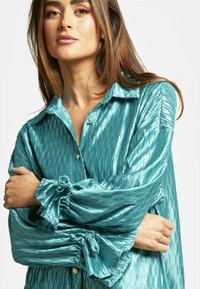 Camisa de satén color verde azulado con un patrón de ondas texturizadas, que presenta una parte delantera con botones, mangas largas y puños fruncidos con detalles de lazo.