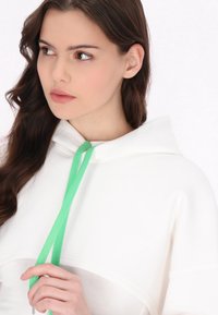 Biała bluza z kapturem i zielonym sznurkiem, charakteryzująca się luźnym krojem i miękką fakturą. Wyjątkowe detale to kaptur oraz prosty, czysty design.