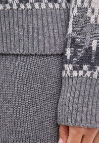 Grauer grob gestrickter Pullover mit geripptem Muster und gestaltetem Design in dunklerem Grau und Creme. Nahaufnahme zeigt detaillierte Nähte und Materialvariationen.