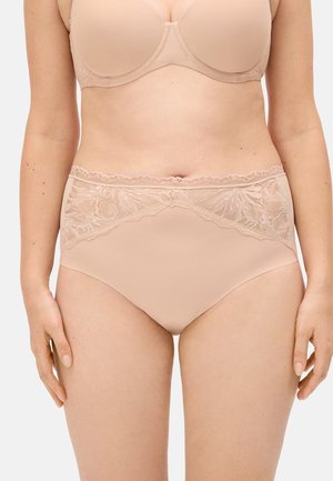 Mujer que lleva ropa interior nude de tiro alto con detalles de encaje y un sujetador a juego, de pie frente a un fondo claro y liso.