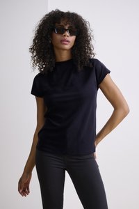 Haut noir à manches courtes avec un col rond, associé à un jean foncé. Le modèle a des cheveux bouclés et porte des lunettes de soleil sombres.