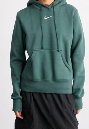 Persona che indossa una felpa con cappuccio verde Nike con tasca frontale e gonna cargo nera, in piedi davanti a uno sfondo chiaro e uniforme.
