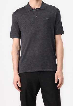 Mężczyzna w ciemnoszarej polo z krótkim rękawem z dwoma guzikami i czarnych spodniach, stojący na tle jednolitego białego tła.