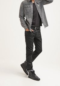 Camisa de denim negra con dos bolsillos en el pecho, superpuesta sobre una camiseta negra. Pantalones ajustados negros y botas altas negras completan el conjunto.