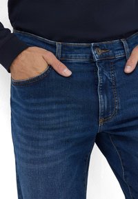 Jean en denim bleu foncé avec un design classique à cinq poches. Les caractéristiques incluent des coutures visibles et une fermeture à bouton à la taille.