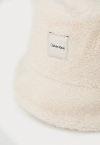 Kremový klobouk typu bucket vyrobený z měkkého, texturovaného fleecu s čtvercovým značkovým štítkem s nápisem "Calvin Klein" po straně.