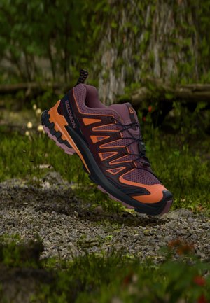 Trail løbesko i lilla og orange farver, svævende over en stenet, mosdækket skovbund.