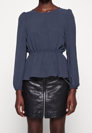 Woman wearing a navy long-sleeve peplum top paired with a black leather mini skirt featuring a front zipper.