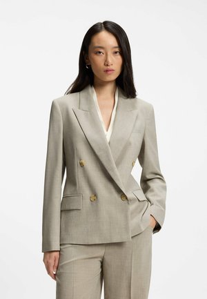 Junge Frau mit langen dunklen Haaren trägt einen beigen zweireihigen Blazer und passende Hose, steht mit einer Hand in der Tasche vor einfachem Hintergrund.