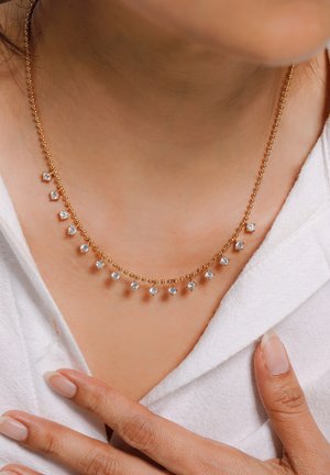 Gold beaded necklace with evenly spaced small diamonds worn on neck, hand with manicured nails adjusting white textured fabric.