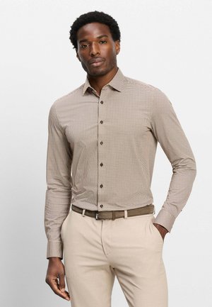 Mann mit kurzen lockigen Haaren trägt ein beiges kariertes Langarmhemd, das in hellbeige Hosen mit braunem Gürtel gesteckt ist, steht mit einer Hand in der Tasche.