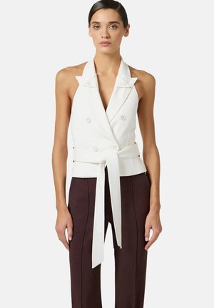 Donna che indossa un blazer bianco smanicato doppiopetto con cintura annodata e pantaloni scuri a vita alta, in piedi davanti a uno sfondo semplice.