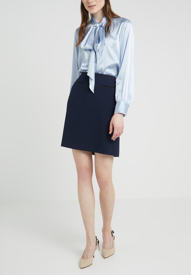 Light blue satin blouse with a tied neckline, long sleeves; navy textured skort with two black accents; beige heels with a bow detail.