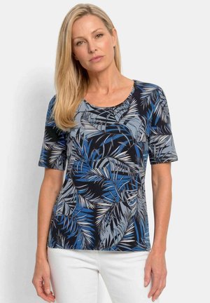 Femme blonde portant une chemise à manches courtes avec un motif de feuilles tropicales bleues, grises et noires et un pantalon blanc, debout sur un fond uni.