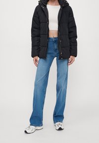 Svart puffjacka med huva och framdragkedja, kombinerad med ljusblå utsvängda jeans och vita sneakers. Har guldaccents.