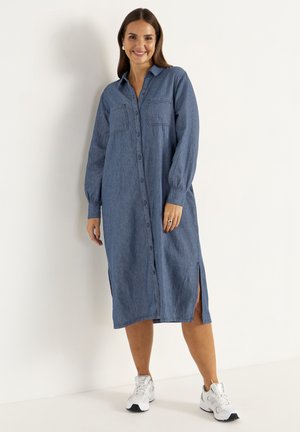 Lichtblauwe denim jurk met lange mouwen, knoopsluiting, twee borstzakken en zijsplitten. Gecombineerd met witte sportschoenen.