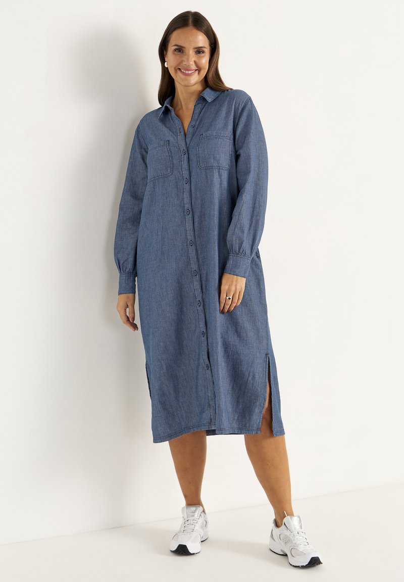 Denim-paita-mekko vaaleansinisessä värissä, jossa on pitkät hihat, nappi edessä, kaksi rintataskua ja sivuhalkiot. Yhdistetty valkoisiin urheilukenkiin.