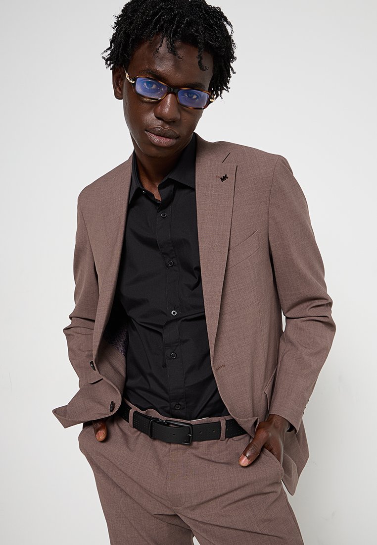 Completo marrone con texture, accompagnato da una camicia nera, caratterizzato da una vestibilità slim, rever a punta e un sottile accento del logo sul rever. Cintura nera e occhiali.