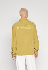 Camicia di velluto a coste gialla senape con vestibilità comoda, caratterizzata da un logo ricamato prominente sul retro e polsini con bottoni.