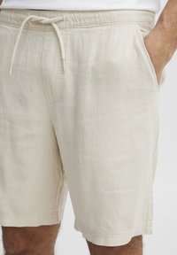 Beige vászon rövidnadrág, elasztikus derékkal és zsinórral. Két oldalsó zseb, egyenes lábszár kialakítás, és látható varrási részletek.